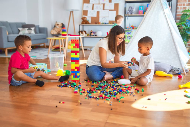 Jovem e bela professora e crianças brincando com blocos de construção em torno de lotes de brinquedos no jardim de infância
 - Foto, Imagem