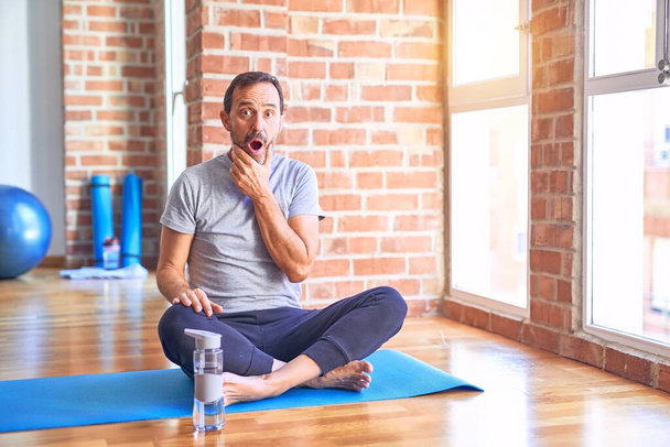 Moyen âge beau sportif assis sur le tapis faire des exercices de yoga étirement à la salle de gym regard fasciné par l'incrédulité, surprise et expression étonnante avec les mains sur le menton
 - Photo, image