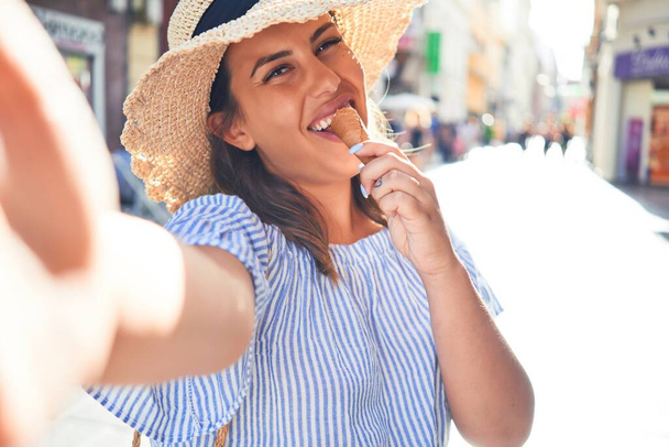 Young beautiful woman eating ice cream cone taking a selfie using smartphone on a sunny day of summer on holidays - Photo, Image
