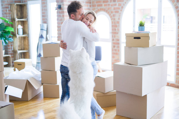 Young beautiful couple with dog hugging at new home around cardboard boxes - Photo, Image