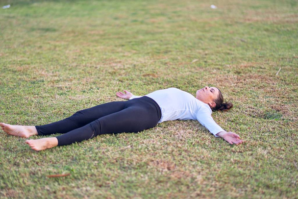 Young beautiful sportwoman practicing yoga. Coach lying down teaching corpose pose at park - 写真・画像