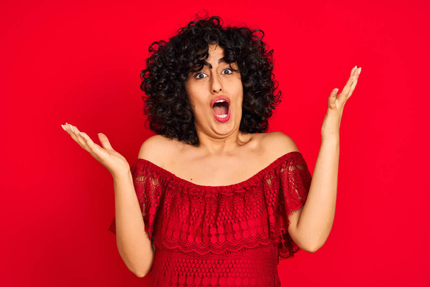 Young arab woman with curly hair wearing casual dress over isolated red background celebrating crazy and amazed for success with arms raised and open eyes screaming excited. Winner concept - Photo, Image