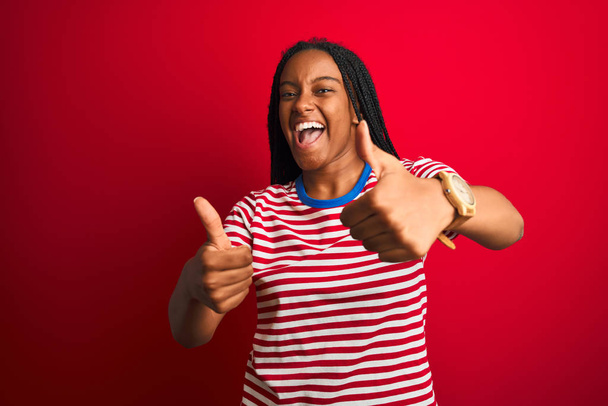Молода афроамериканська жінка, одягнена в смугасту футболку, стоїть над ізольованим червоним фоном, схвалюючи позитивний жест рукою, великий палець піднімає посмішку і щасливий за успіх. Шедевр переможця.. - Фото, зображення