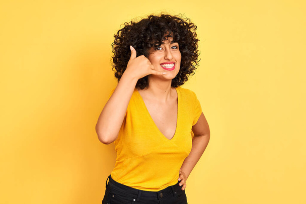 Jeune femme arabe aux cheveux bouclés portant un t-shirt debout sur fond jaune isolé souriant faisant geste téléphonique avec la main et les doigts comme parler au téléphone. Communication des concepts
. - Photo, image