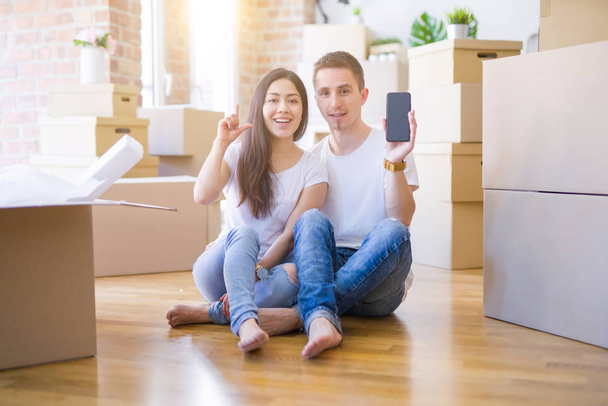 Hermosa pareja sentada en el piso sosteniendo el teléfono inteligente en el nuevo hogar alrededor de cajas de cartón sorprendida con una idea o pregunta apuntando con el dedo con la cara feliz, número uno
 - Foto, imagen