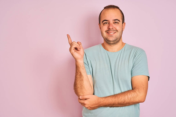 Hombre joven con una camiseta casual azul de pie sobre un fondo rosa aislado con una gran sonrisa en la cara, apuntando con la mano y el dedo hacia el lado mirando a la cámara
. - Foto, imagen