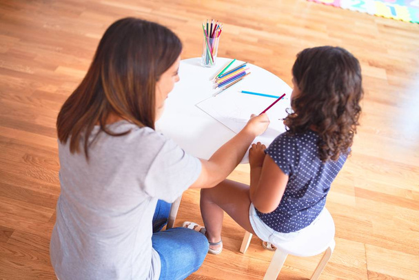 Mooie leraar en peuter meisje tekening tekenen met behulp van gekleurde potloden op de kleuterschool - Foto, afbeelding