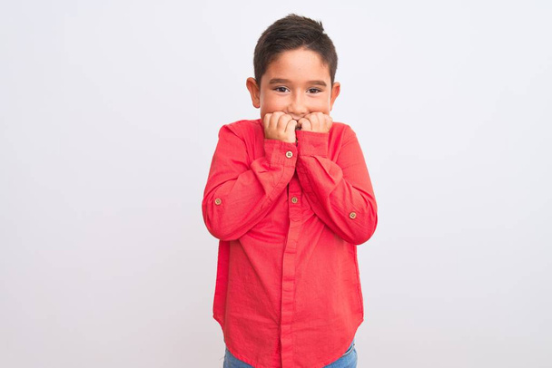 孤立した白い背景の上に立つエレガントな赤いシャツを身に着けている美しい少年は、口の上に爪をかむ手で強調し、緊張している。不安の問題. - 写真・画像