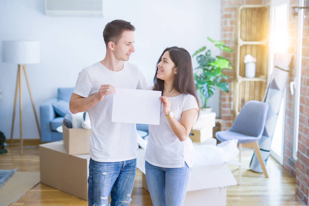 Young beautiful couple holding banner at new home around cardboard boxes - Foto, Bild