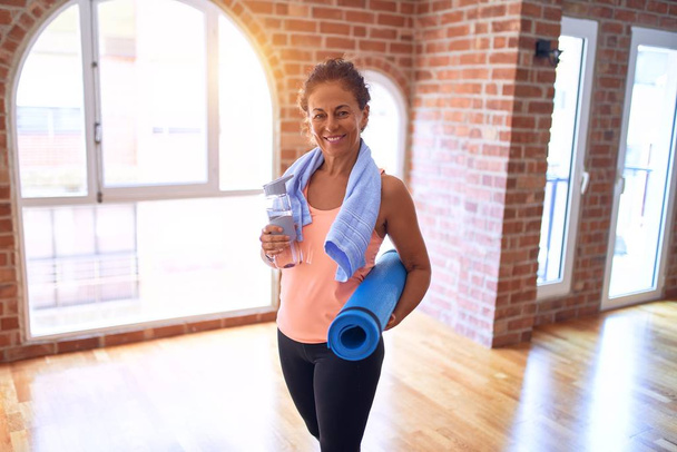 Middle age beautiful sportwoman smiling happy and confident. Standing with smile on face holding mat and bottle of water before doing exercise at gym - Фото, зображення