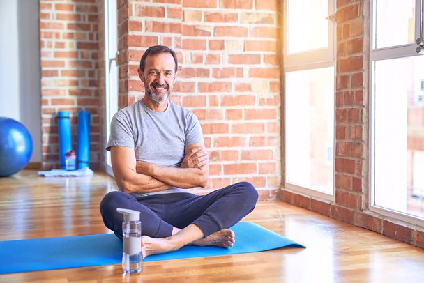 Middelbare leeftijd knappe sportman zittend op mat doen stretching yoga oefening op sportschool gelukkig gezicht glimlachen met gekruiste armen kijken naar de camera. Positief persoon. - Foto, afbeelding