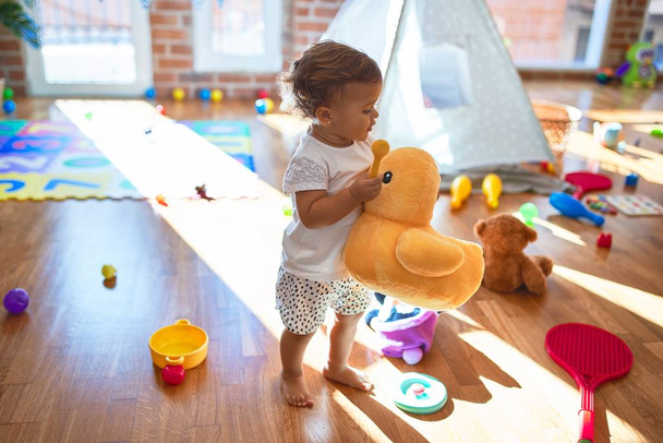 Adorable toddler holding duck doll standing around lots of toys at kindergarten - Foto, afbeelding