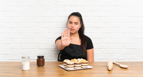 Joven adolescente asiática chica con un montón de pastel de magdalenas haciendo stop gesto con su mano
 - Foto, Imagen