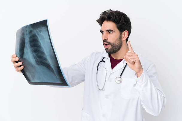Giovane bell'uomo con la barba su sfondo bianco isolato indossando un abito da medico e tenendo una scansione ossea - Foto, immagini
