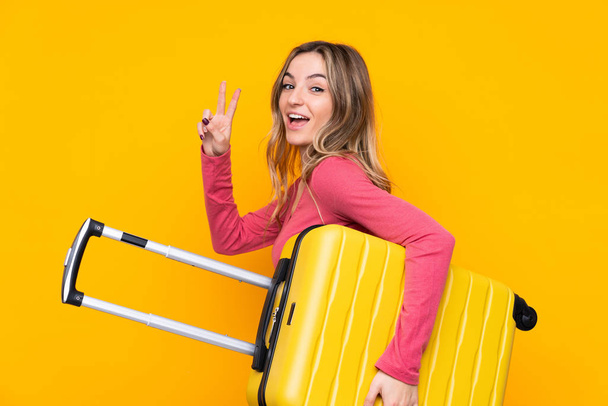 Young woman over isolated yellow background in vacation with travel suitcase and making victory gesture - Foto, Imagem