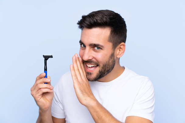 Young handsome man shaving his beard over isolated background whispering something - Photo, Image