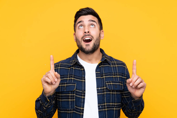 Young handsome man with beard over isolated yellow background surprised and pointing up - 写真・画像