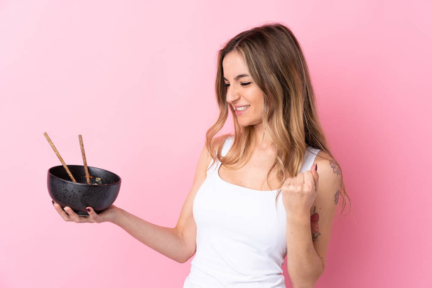Young woman over isolated pink background celebrating a victory while holding a bowl of noodles with chopsticks - Фото, изображение