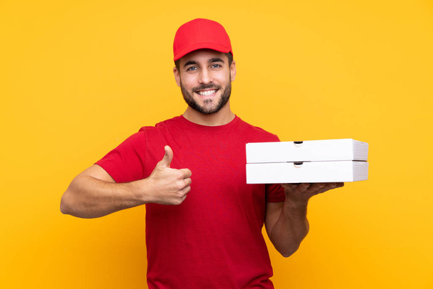 pizza delivery man with work uniform picking up pizza boxes over isolated yellow background with thumbs up because something good has happened - Foto, Imagen