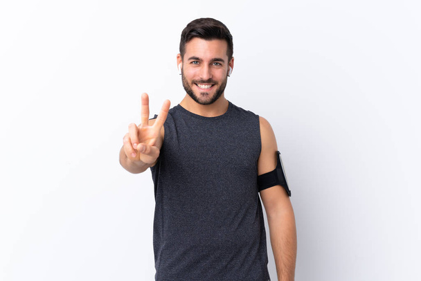 Mladý sport pohledný muž s vousem přes izolované bílé pozadí s úsměvem a ukazující vítězství znamení - Fotografie, Obrázek