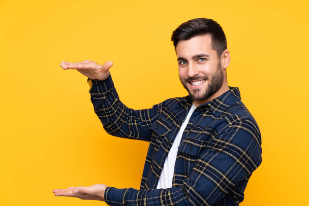 Joven hombre guapo con barba sobre fondo amarillo aislado sosteniendo espacio para insertar un anuncio
 - Foto, imagen