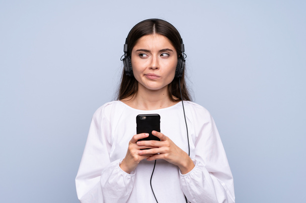 junge Frau über einsamen blauen Hintergrund mit dem Handy mit Kopfhörer und Denken - Foto, Bild