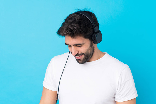 Junger gutaussehender Mann mit Bart vor isoliertem blauen Hintergrund, der Musik hört - Foto, Bild
