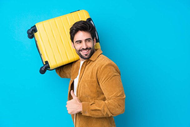 Νεαρός όμορφος άνδρας με γενειάδα πάνω από απομονωμένο μπλε φόντο σε διακοπές με ταξιδιωτική βαλίτσα και με τον αντίχειρα επάνω - Φωτογραφία, εικόνα