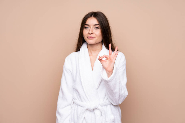 Молодая девушка в халате на изолированном фоне показывает знак ОК с пальцами - Фото, изображение