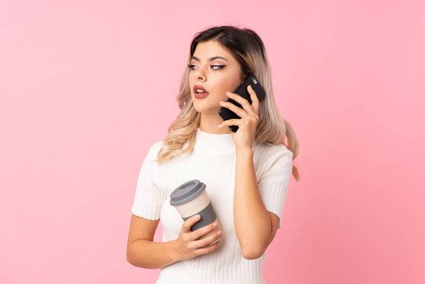 Teenager girl over isolated pink background holding coffee to take away and a mobile - Фото, зображення