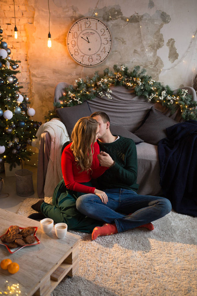 a happy, loving couple is sitting near the sofa, next to it is a Christmas tree, a table and tangerines, cups with chocolate, coffee with milk, cocoa, hugs. tenderness and comfort in the house. - Photo, Image