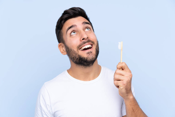 Young handsome man with beard brushing his teeth over isolated background looking up while smiling - Foto, Bild