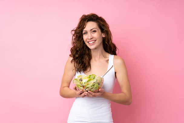 Jonge vrouw met krullend haar met een salade over geïsoleerde roze achtergrond - Foto, afbeelding