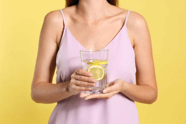 Young woman with glass of lemon water on yellow background, clos - Foto, afbeelding