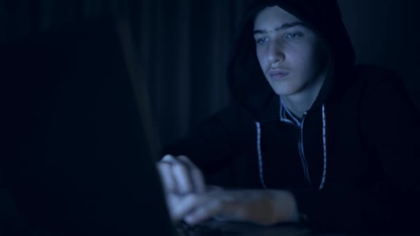 Hacker's hacking into the computer network. A young guy in a dark room typing on a laptop.  Hacker with a laptop. E-the robber. Lonely teen chatting online. Computer criminal. 4K footage - Metraje, vídeo