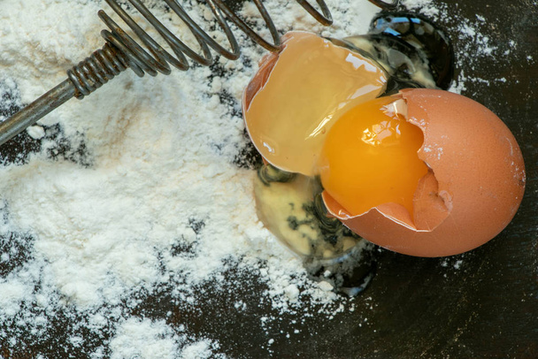 Mutfak masasında buğday unu, kırık yumurta ve çırpmak için çırpılmış çırpma. - Fotoğraf, Görsel