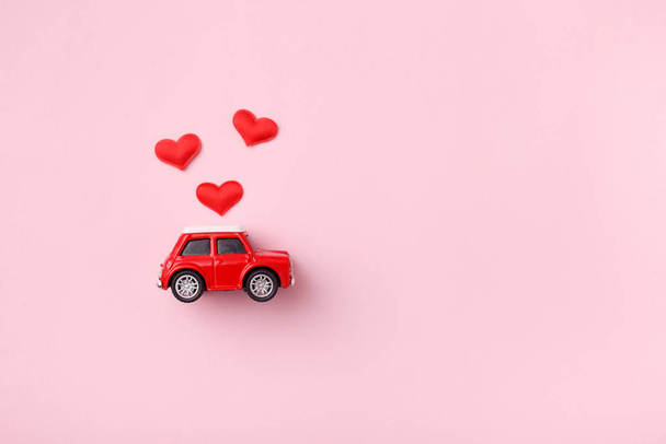 Red retro toy red car with red bow for Valentine's day on pink background with heart confetti. Top view, flat lay - Foto, Imagen