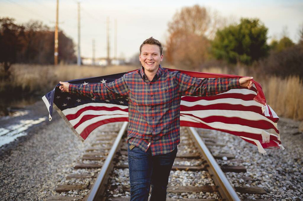 Smiling male holding the United States flag while walking on the train rails - Foto, afbeelding