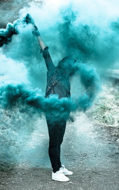 Vertical shot of a person making a blue-colored smoke with a smoke bomb while standing on the road - Foto, Imagem