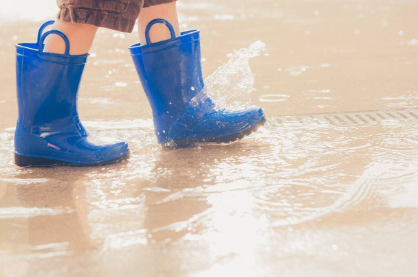 Closeup shot of the legs of a boy in blue rubber boots splashing in a puddle - Фото, зображення