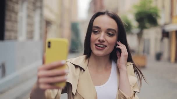 Beautiful Smiling Young Lady Wearing Earphones touching them Making a Video call Holding Phone Vertical Talking and Standing on the City Street Background Close Up. - Materiaali, video