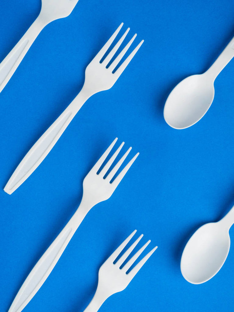 Πλαστικό λευκό πιρούνι μιας χρήσης και κουτάλι σε ένα μοντέρνο μπλε χρώμα φόντο. Έννοια πλαστικά πιάτα, fast food, πλαστική ρύπανση. Πάνω άποψη, επίπεδη lay. - Φωτογραφία, εικόνα