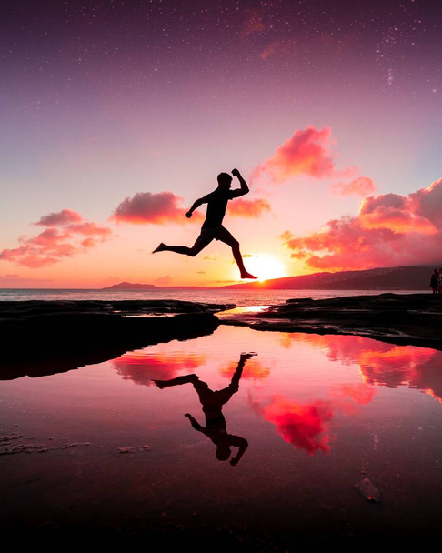 A vertical shot of a person jumping over the water and his reflection in it under the pink sunset sky - Foto, Imagen