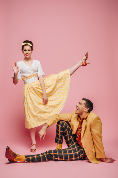 cheerful man sitting on floor and girl waving at camera while dancing boogie-woogie on pink background - Photo, Image