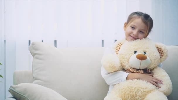 Happy little girl sitting on the sofa, hugging a big teddy-bear. - Πλάνα, βίντεο