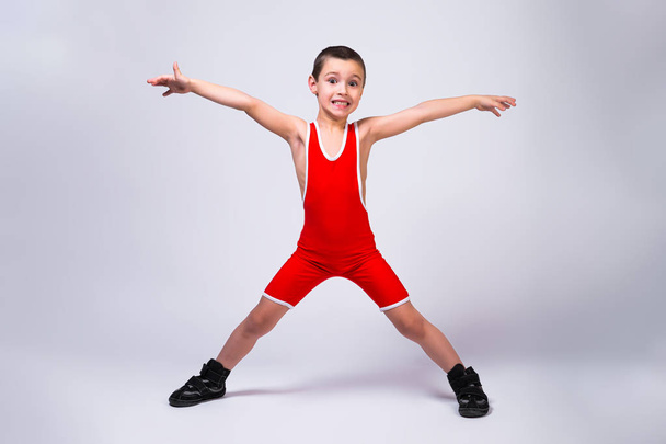 Malý zábavný chlapec v červeném sportovním ortsovskoy trikotu a wrestling boty drží ruce a nohy v různých směrech a usmívá se na jeho úspěchy na bílém izolovaném pozadí. Koncept malého sportovce v boji.  - Fotografie, Obrázek