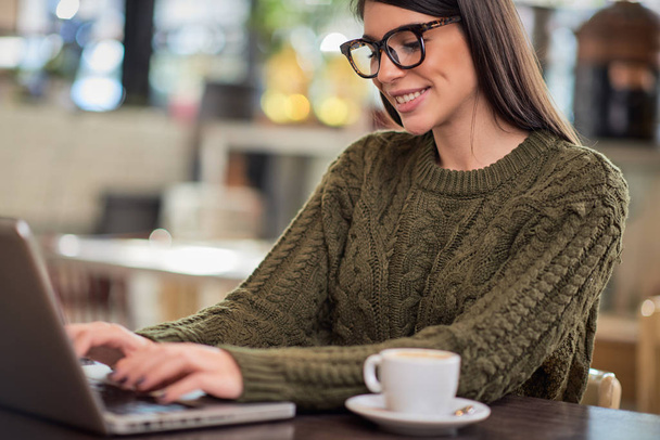 Uśmiechnięta biała brunetka w okularach ubrana swobodnie, siedząca w kawiarni i pracująca na laptopie. Ręce na klawiaturze.. - Zdjęcie, obraz