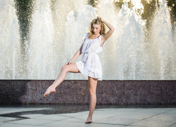 Attractive girl in white short dress sitting in front of a fountain in the summer hottest day. Girl with dress partly wet dancing. Beautiful blonde women near the fountain in a ballet position - Fotoğraf, Görsel