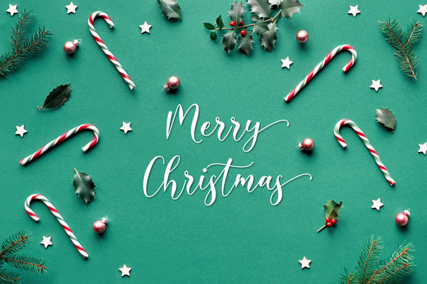 Trendy geometric Christmas flat lay on green paper background with candy canes, holly and fir twigs, wooden stars and glass trinkets, greeting text "Merry Christmas". - Fotografie, Obrázek