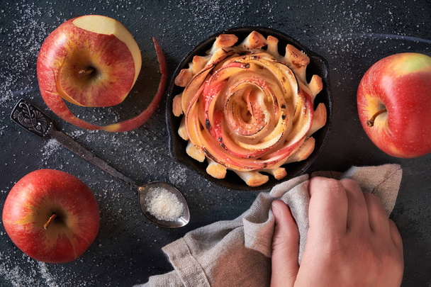 Puff pastry with rose shaped apple slices baked in iron skillet. Top lay on the pie on dark background with some apples and sugar. Hand holding the skillet through the towel. Apple homemade dessert. - Foto, immagini